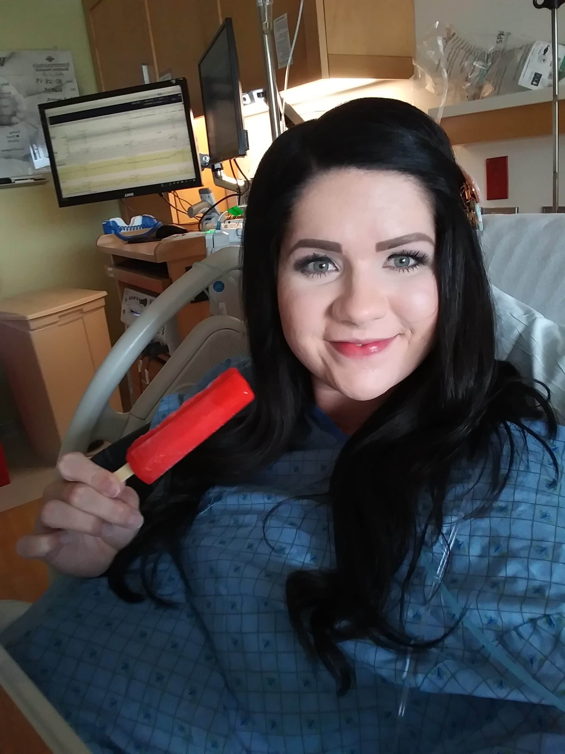 Pregnant brunette woman holds up a popsicle during and takes a selfie in hospital bed.