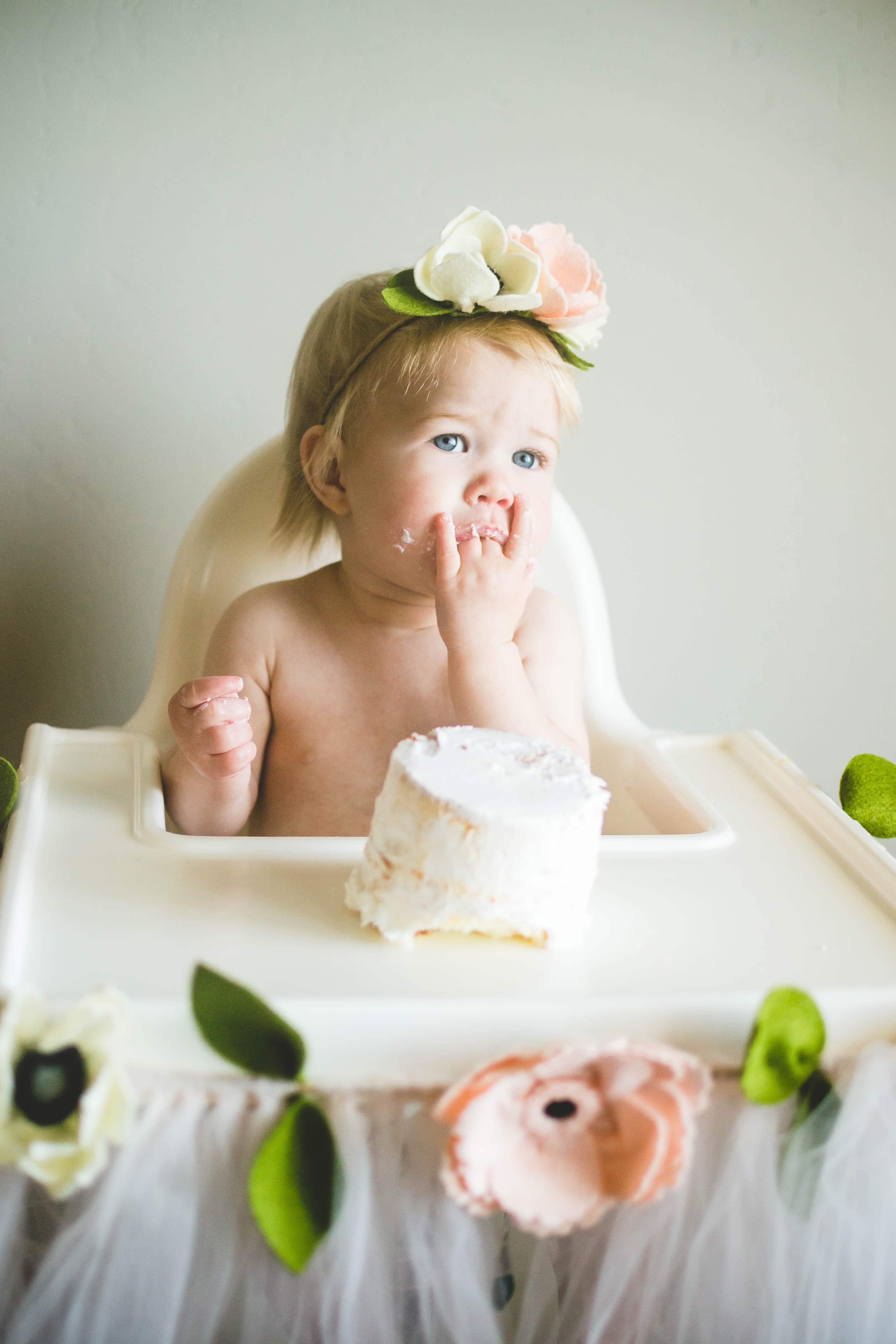 Blonde baby girl wearing floral headband sucks fingers in high chair with birthday cake.