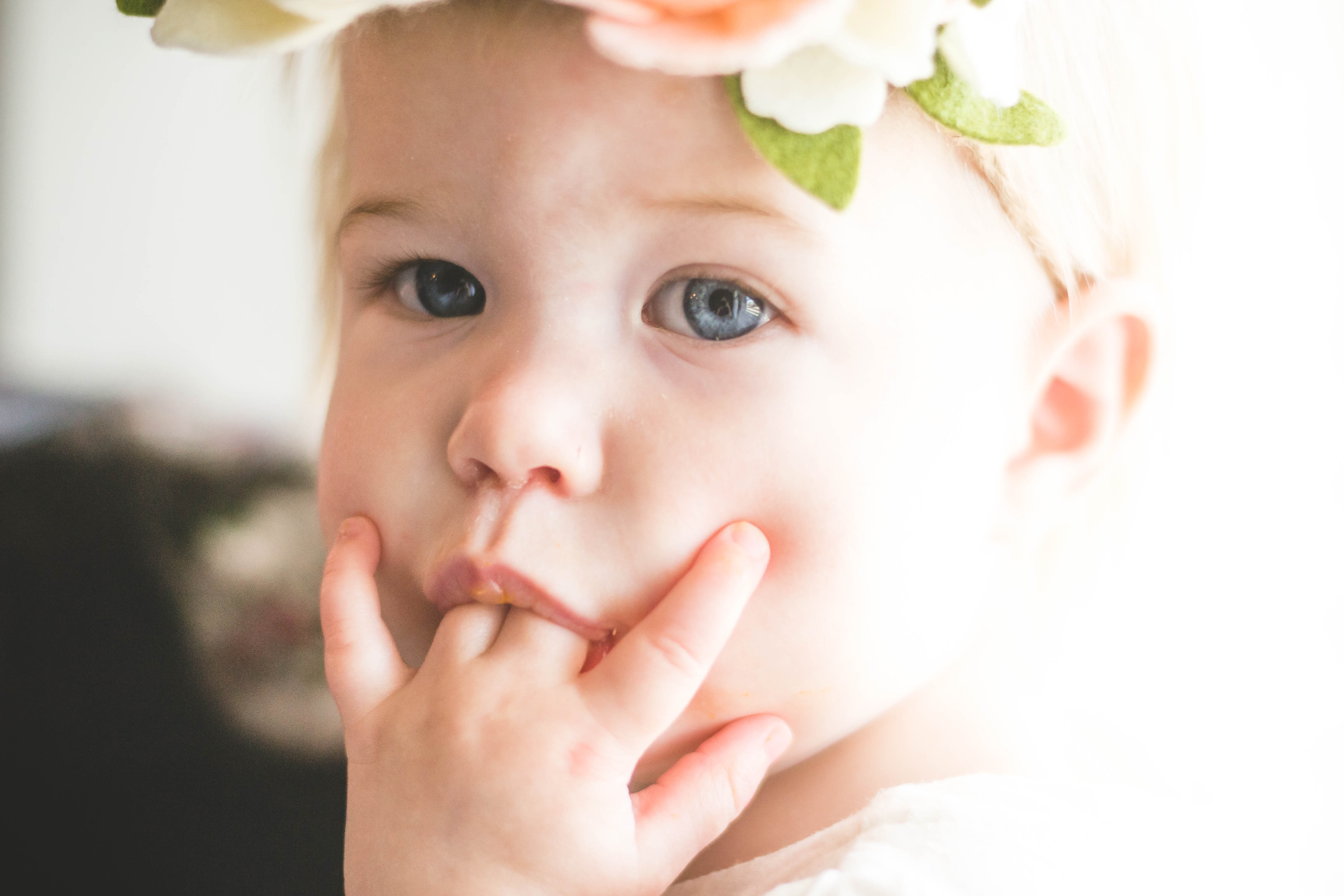 Close up photo of blonde baby girl wearing floral headband sucking fingers.