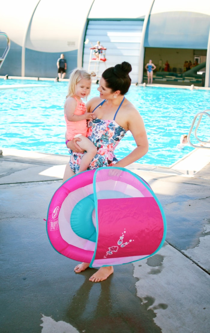 A woman holds her toddler and a pink and blue pool float by the pool.