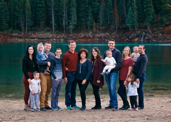 Extended family of 14 wearing jewel tones poses for family picture near lake with pines. 