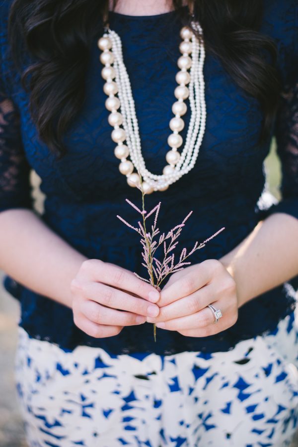 Close up photo of woman wearing blue shirt and pearl necklace holding a pink branch.