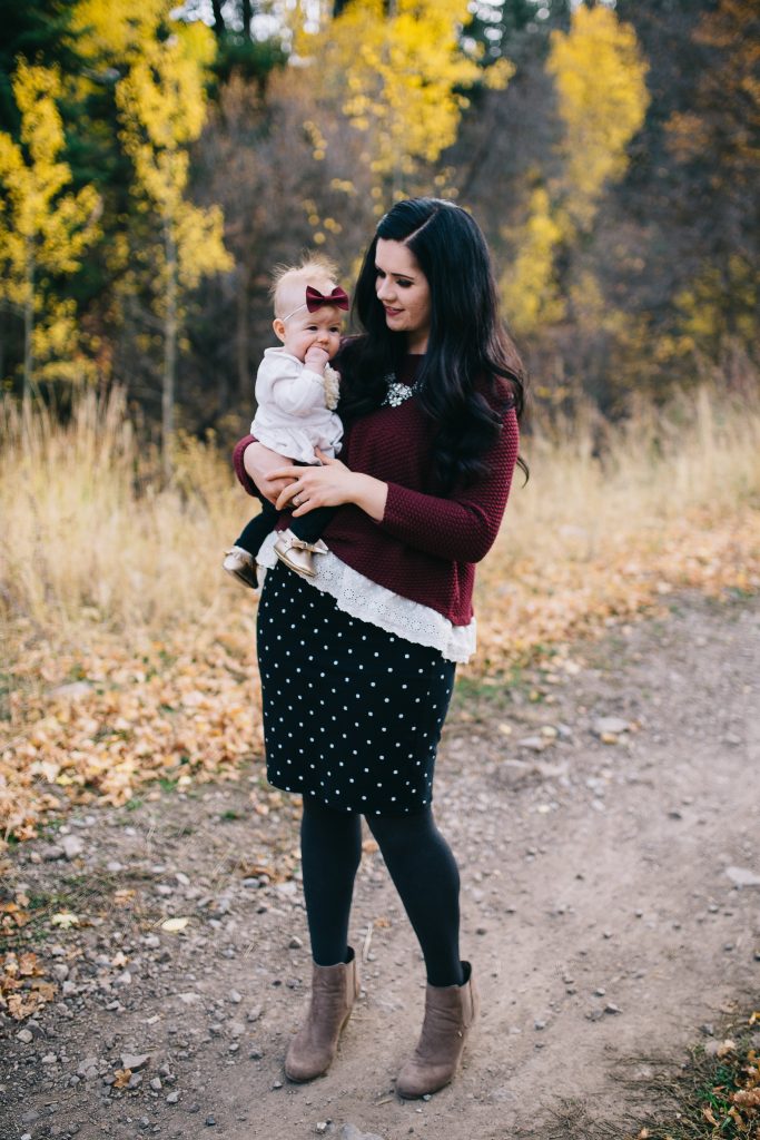 Woman wearing burgundy sweater and black skirt holds baby girl with fist in mouth during fall family pictures.