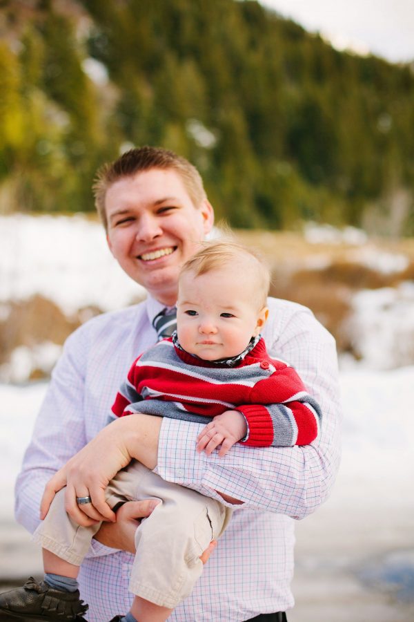 Man wearing red and gray stripe shirt holds baby boy wearing red and gray sweater during mountain photo shoot.