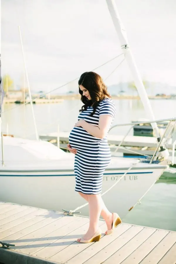 Brunette woman wearing blue stripe dress holds baby belly during maternity photo shoot on dock.