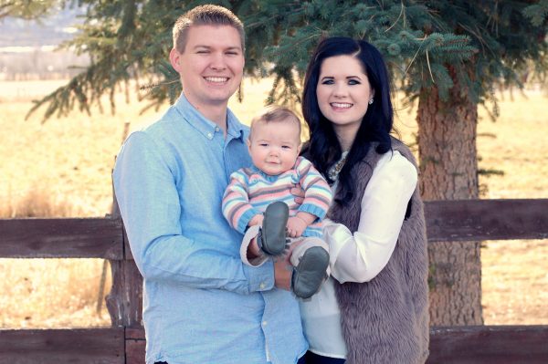 Man wearing blue shirt and woman wearing cream shirt and fur vest hold baby boy and smile during fall family pictures.