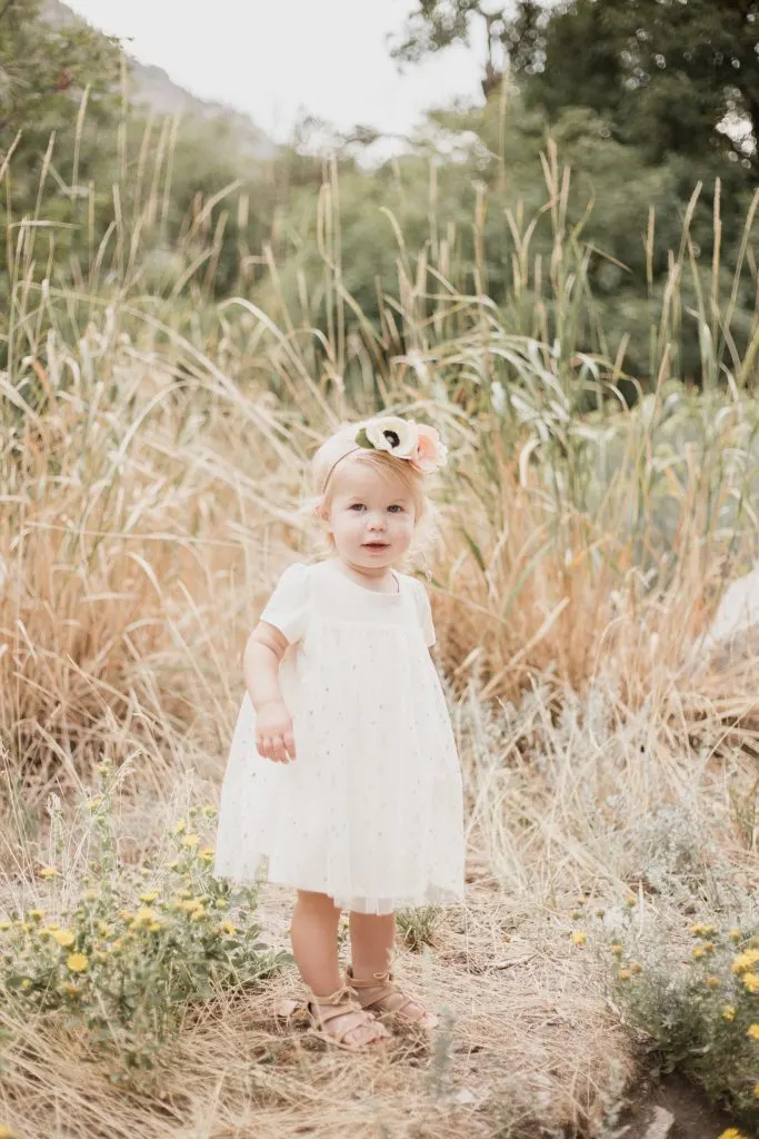 Baby girl wearing cream dress and floral headband stands in grassy field during family pictures.
