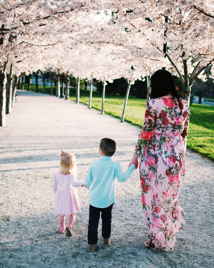 Woman holds hands with two children and walks down gravel path lined with cherry blossom trees.