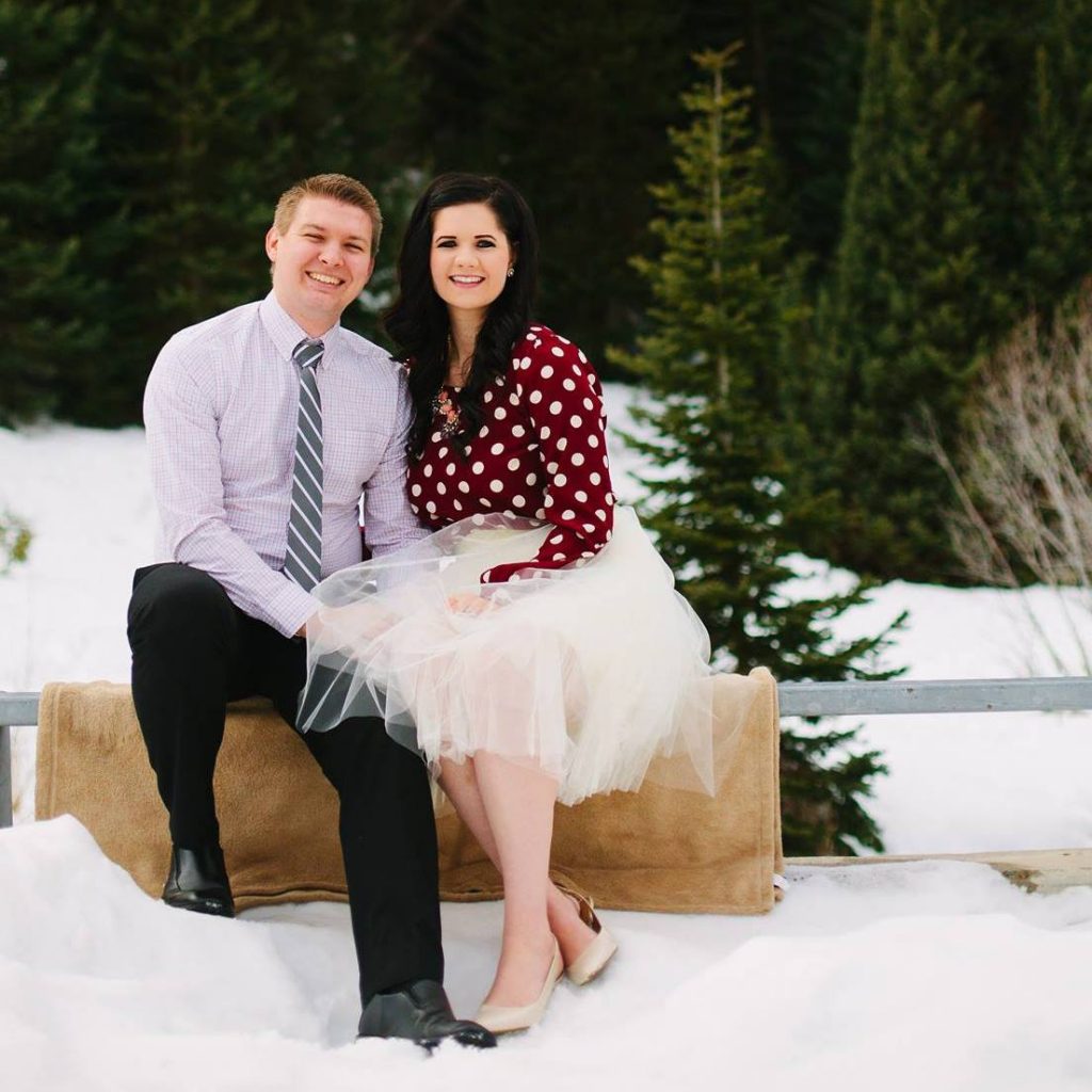 Couple wearing red, cream, and grey outfits sit close on railing and smile during winter photo shoot.