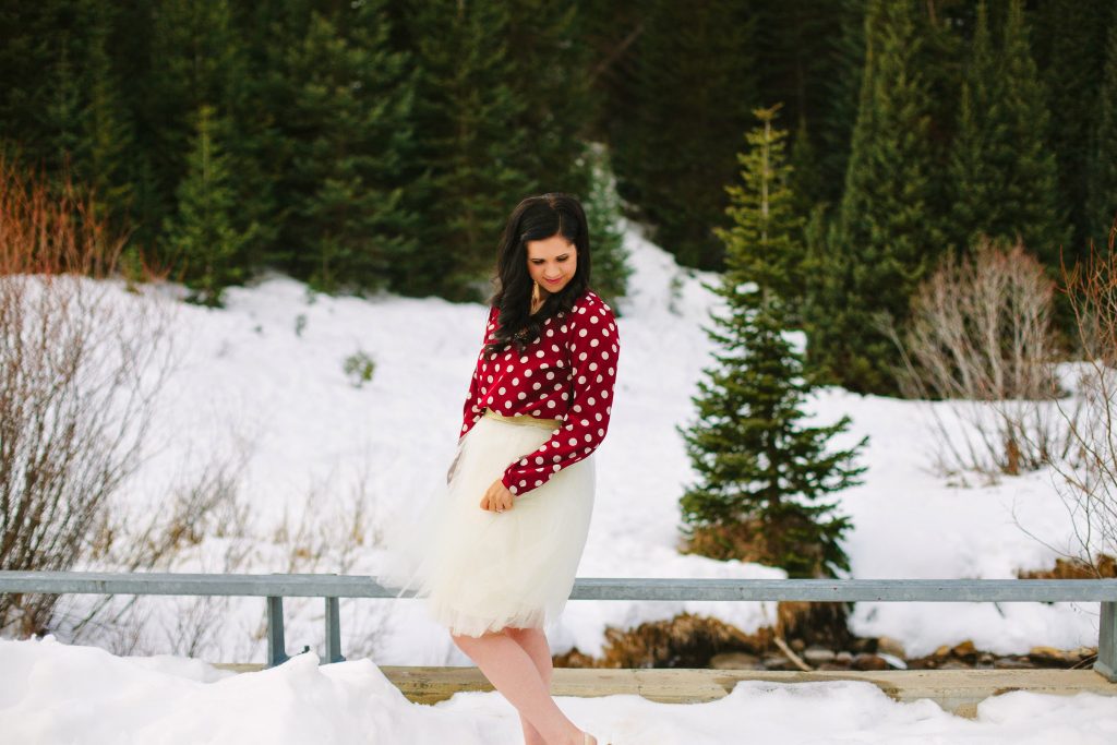 Woman wearing red polka dot shirt holds tulle skirt and looks down during winter photos.