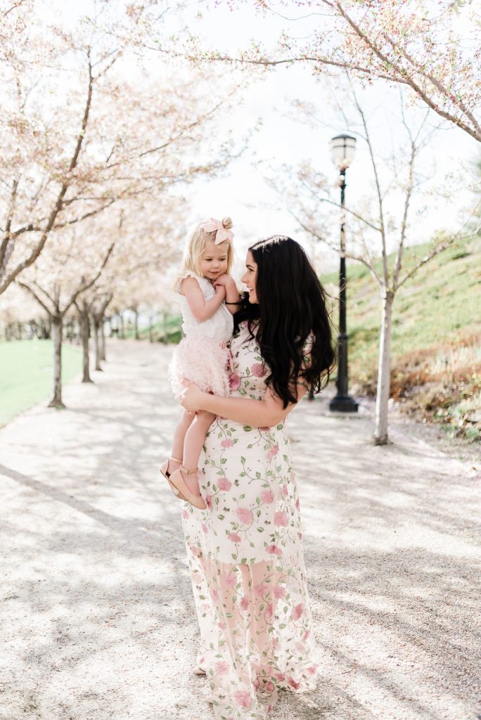 Woman wearing floral dress smiles and holds girl wearing pink skirt and near cherry blossom trees.