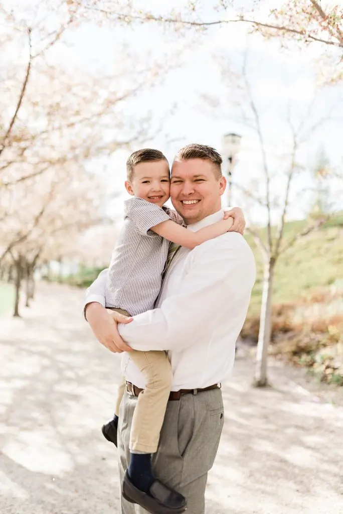 Man wearing white shirt and gray pants holds boy wearing gray shirt as they hug tight during family photos.