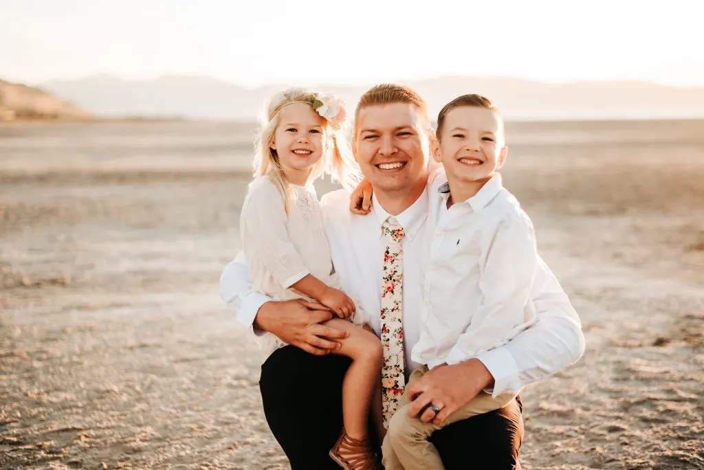 Man holds son and daughter on his legs and smiles during family pictures on beach.