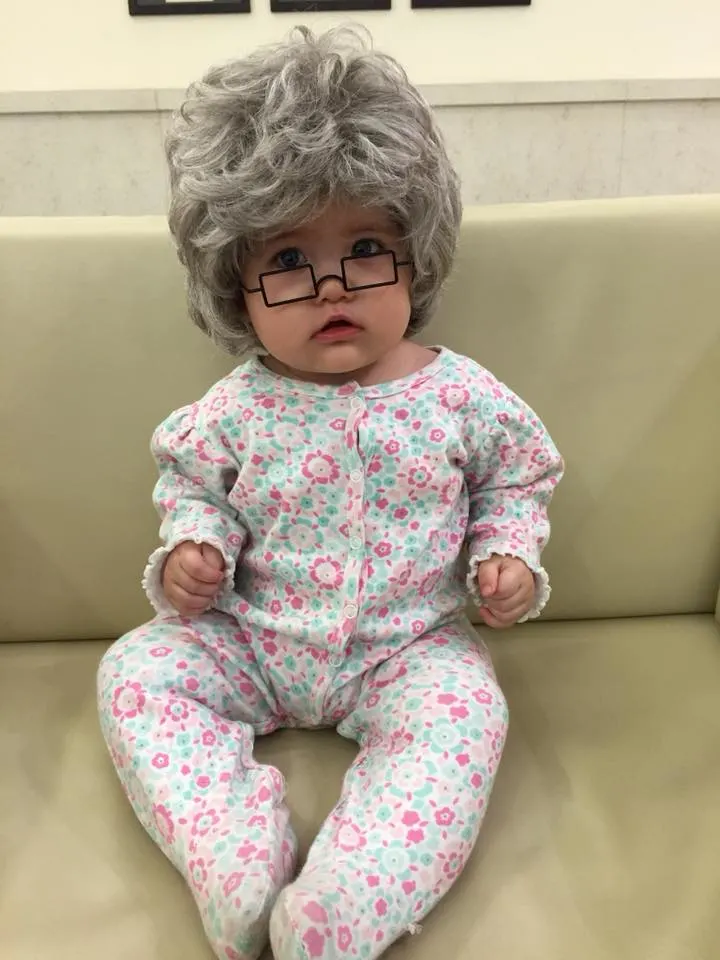 A baby in a floral onesie, a gray wig, and fake glasses sitting on a couch.