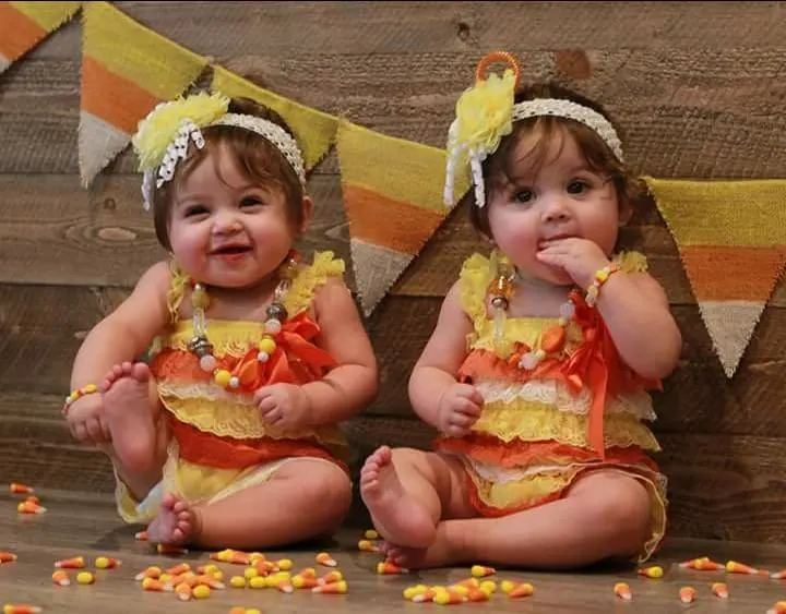 Two twin girls wearing candy corn baby Halloween costumes sit in front of photo backdrop.