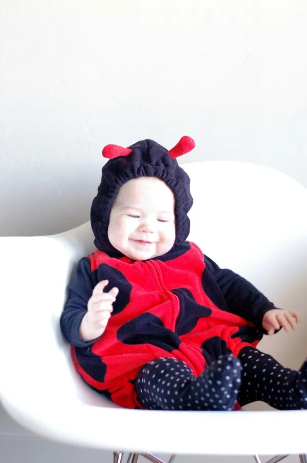 Baby girl smiles while wearing a lady bug Halloween costume and sitting on white chair.