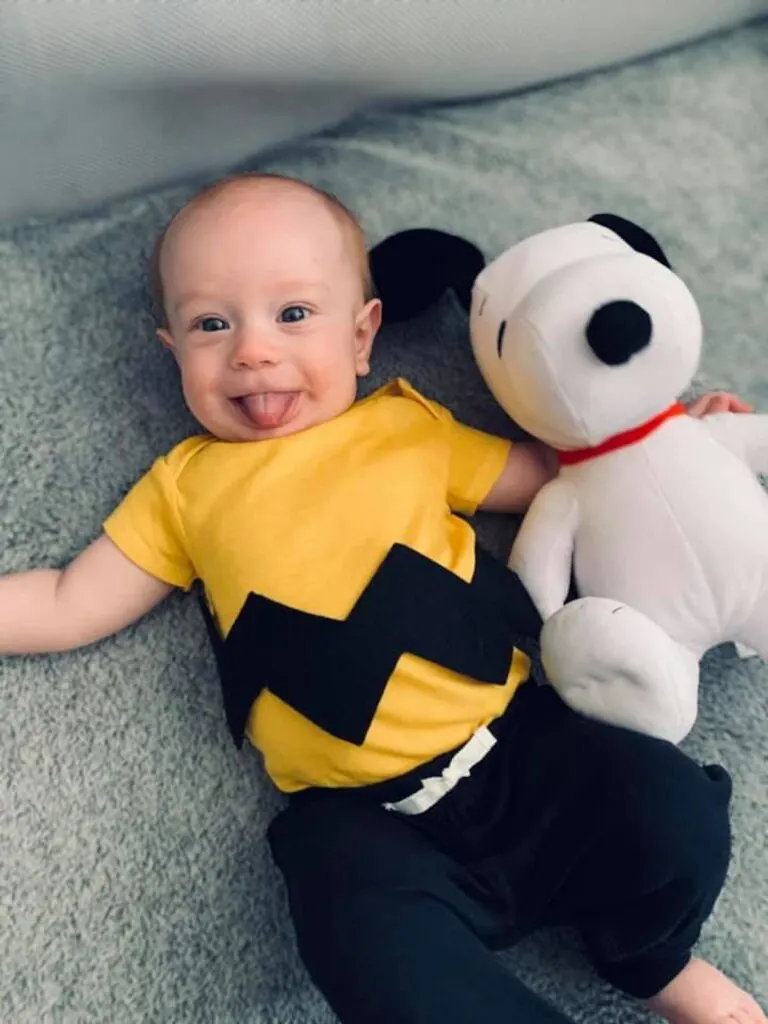 Baby boy wears Charlie Brown costume and lays next to Snoopy dog.