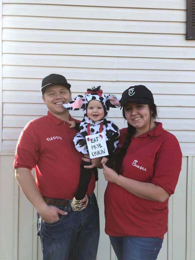 Man and woman wear Chick Fil A shirts and hold baby girl wearing cow costume.