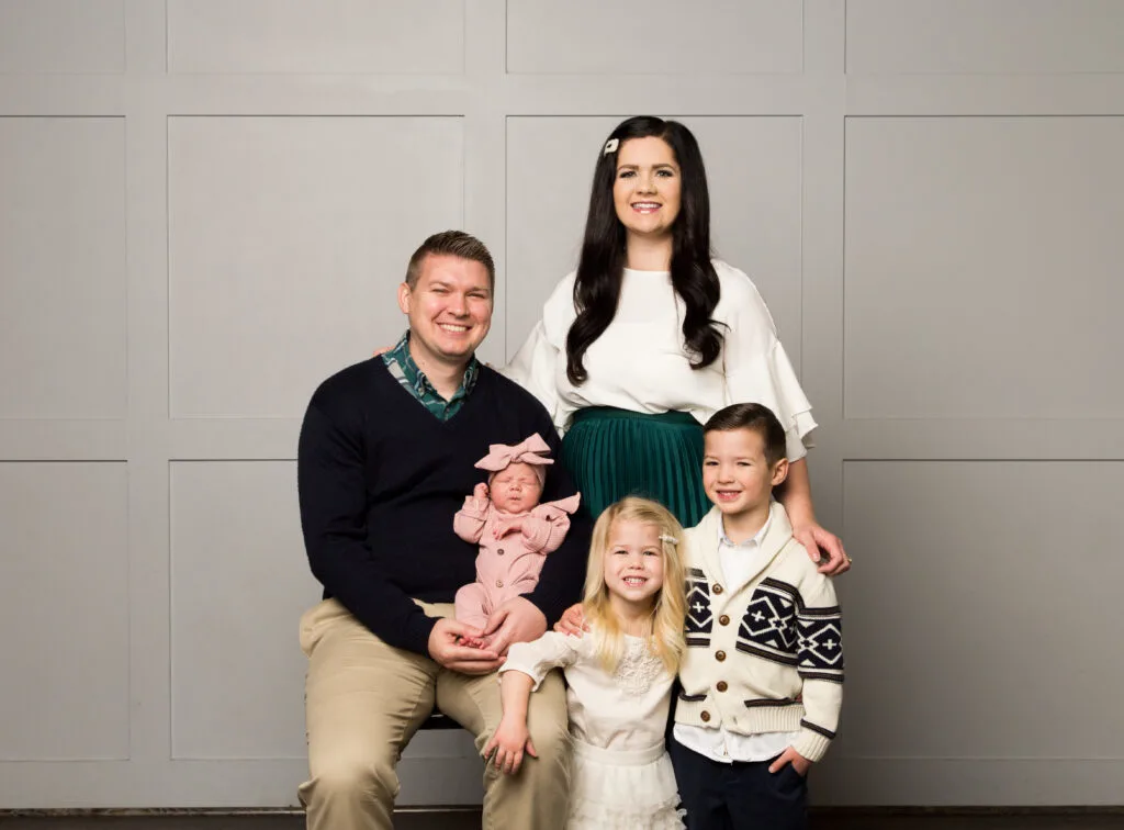 Family of 5 wearing cream, green, and pink smiles for family pictures in front of gray wall.