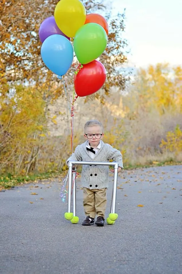 A child dressed up as an old man from the movie "Up" with a walker and holding colorful balloons.