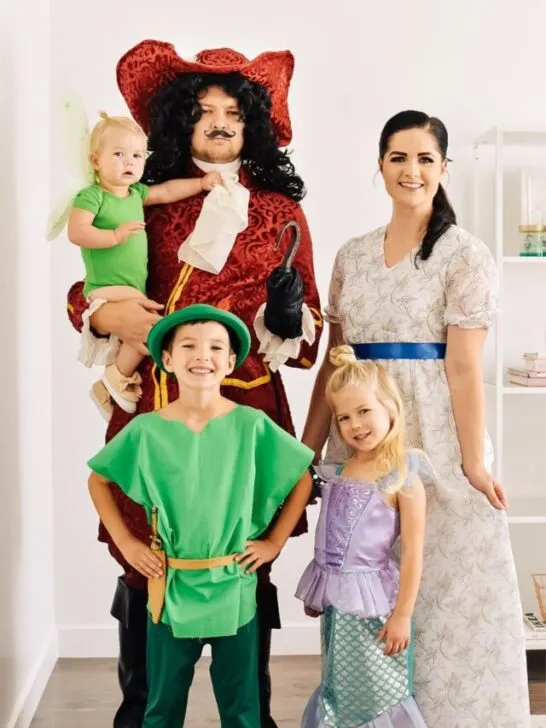 Family wearing Peter Pan character costumes smiles in front of white wall.