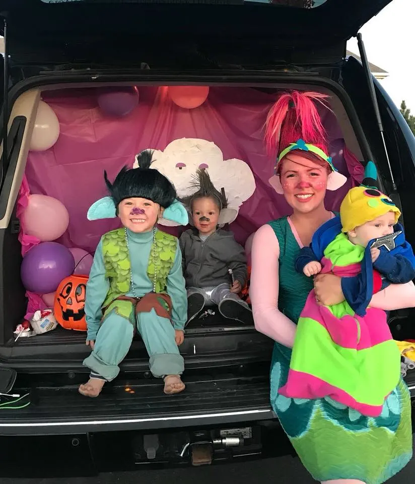Family wearing Trolls Halloween costumes sits in back of decorated car trunk.