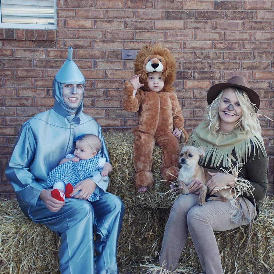 Family wearing Wizard of Oz Halloween costumes sits on bales of hay and smiles.