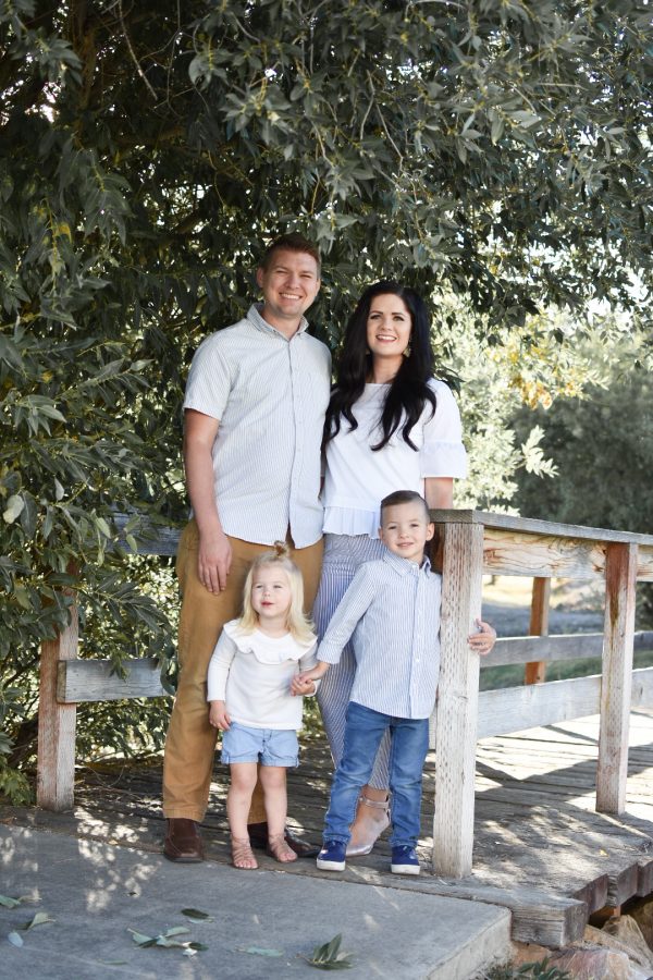 Family wearing cream and blue outfits stands on wood bridge by olive trees and smiles.