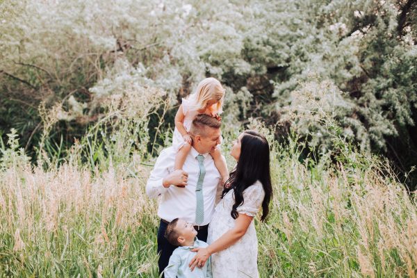 Family stands in grassy field and smiles at each other during summer family photos.
