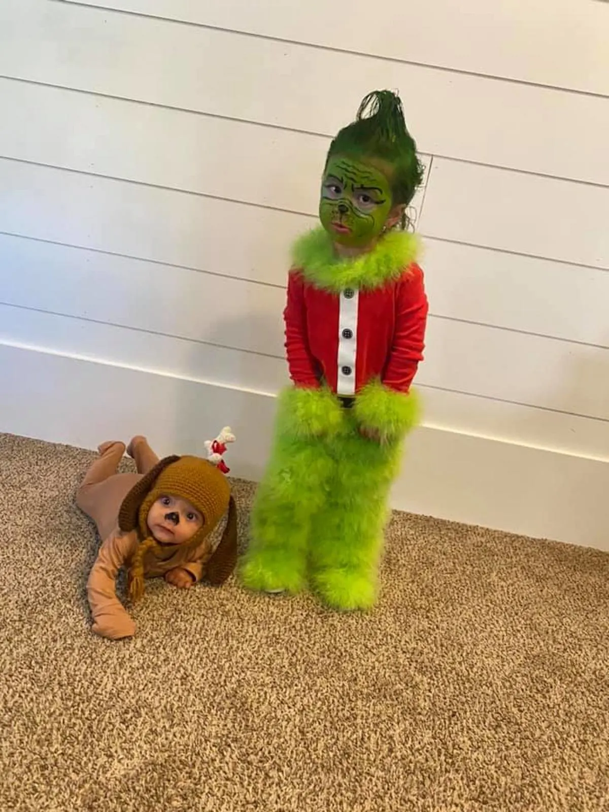 Boy wearing Grinch costume and baby wearing Max costume stand in front of white wall.