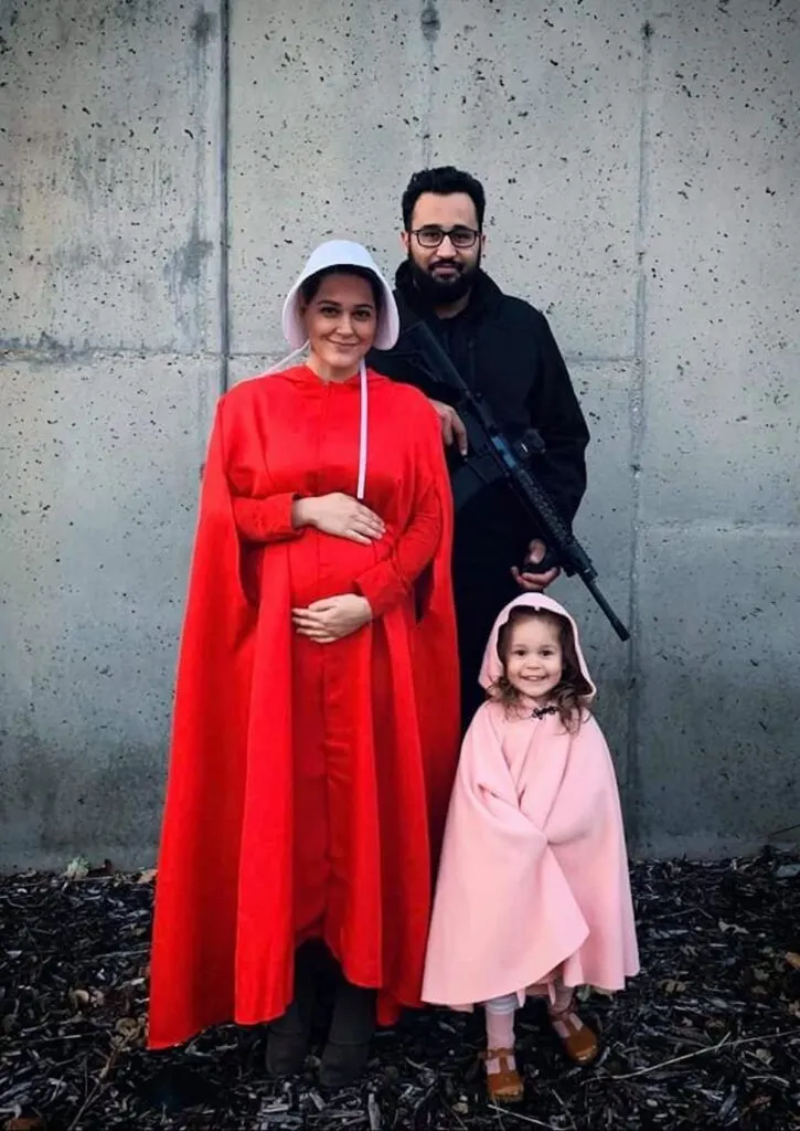 Family dressed as Handmaid's Tale stands in front of cement wall and smiles.