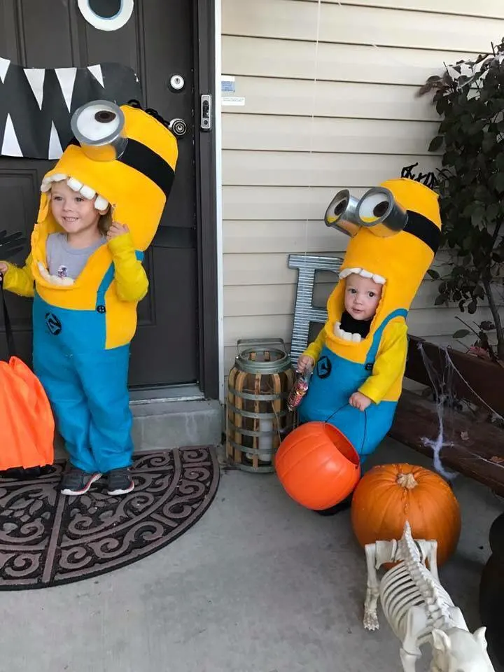 Brother and sister wearing Minion kids Halloween costumes smile on porch with candy buckets.