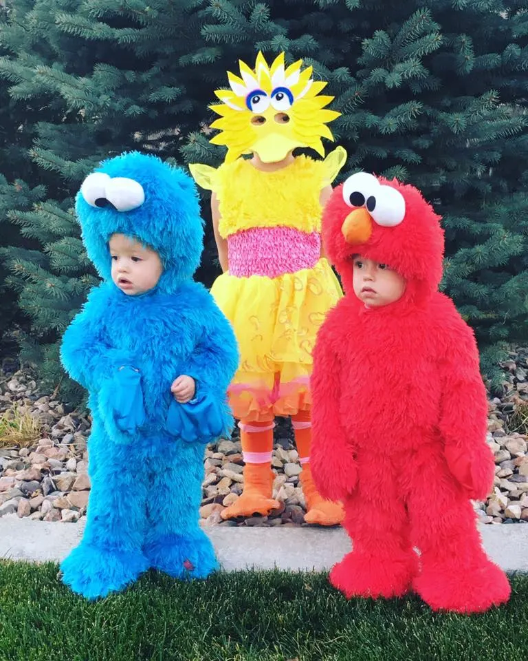 Three siblings wearing Sesame Street kids Halloween costumes stand in front of pine tree.