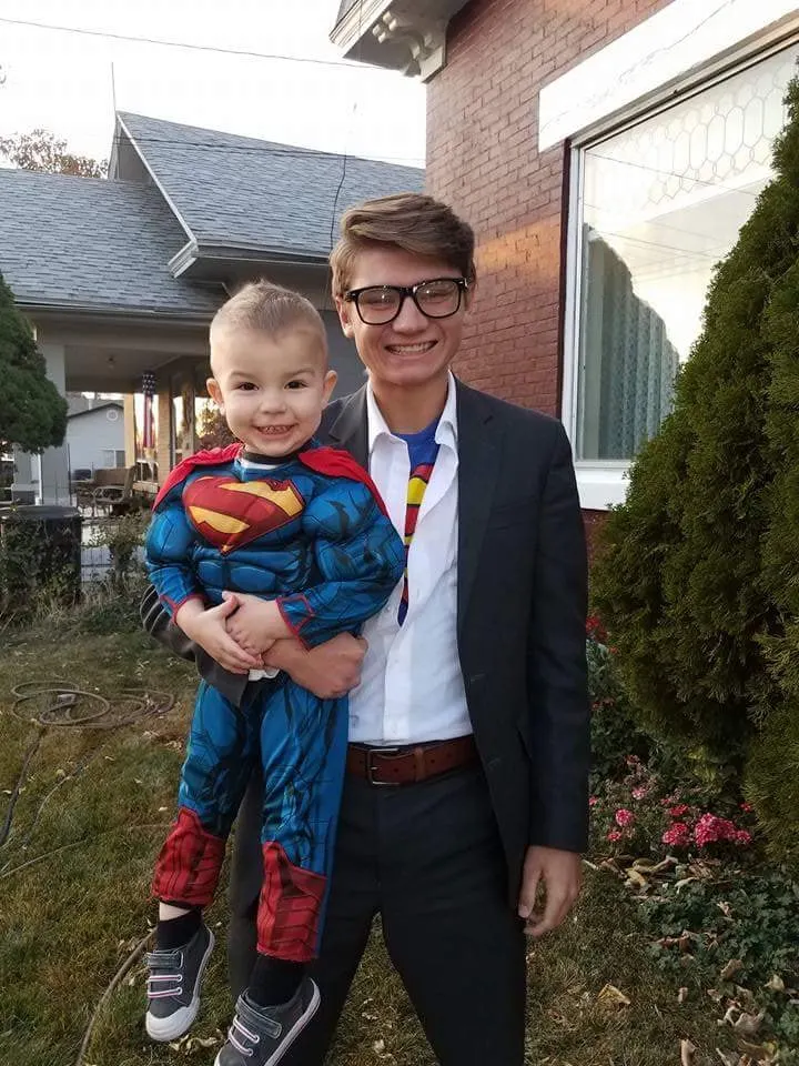 Teenager wearing Clark Kent costume holds baby boy wearing Superman costume.