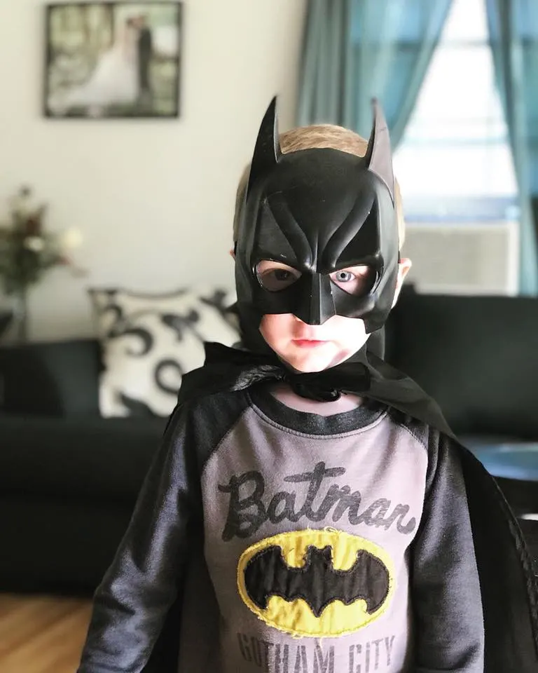 Close up photo of boy wearing Batman kids Halloween costume with living room in background.