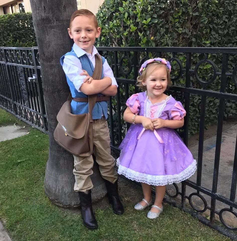 Brother and sister dressed as Rapunzel and Flynn Rider smile next to tree and black gate.