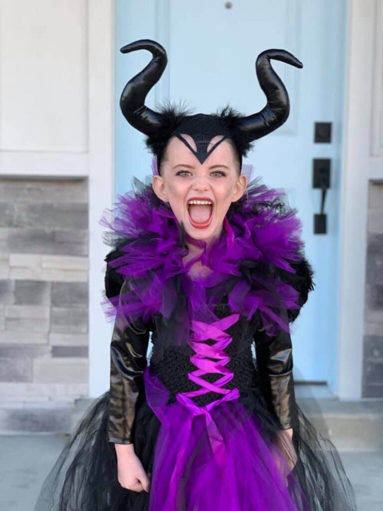 Girl wearing Maleficent costume makes scary face in front of blue door.