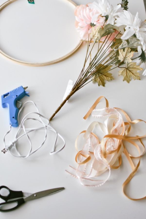 DIY wreath supplies including glue gun, ribbon, wood hoop, and faux flowers on white table.