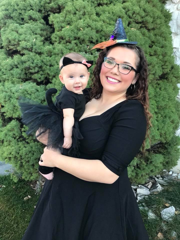 Brunette woman wearing witch costume holds baby girl wearing cat Halloween costume.