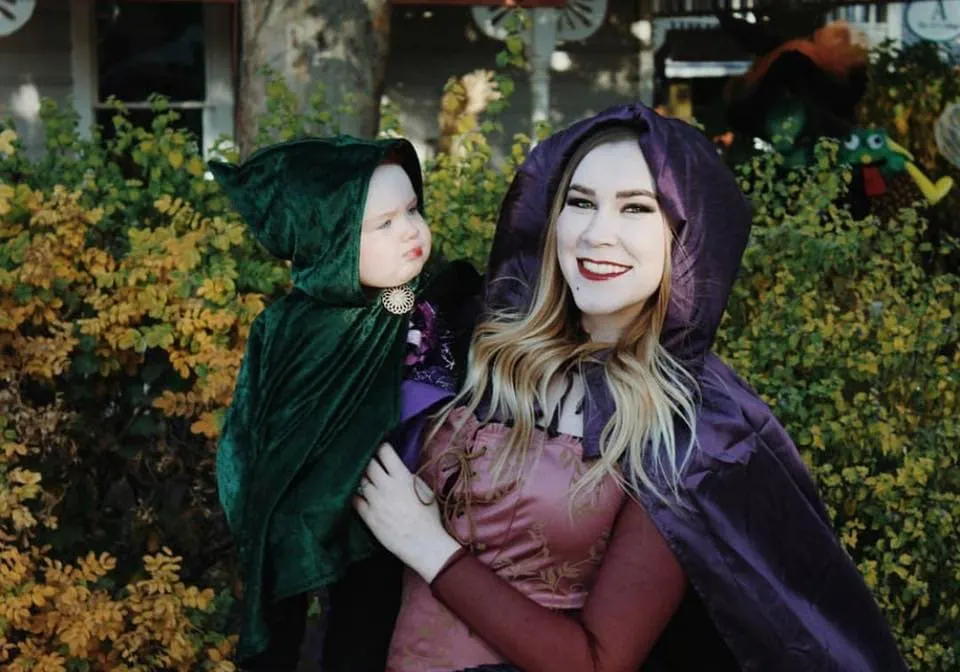 Woman wearing Sarah from Hocus Pocus costume holds baby girl wearing Winifred Halloween costume.