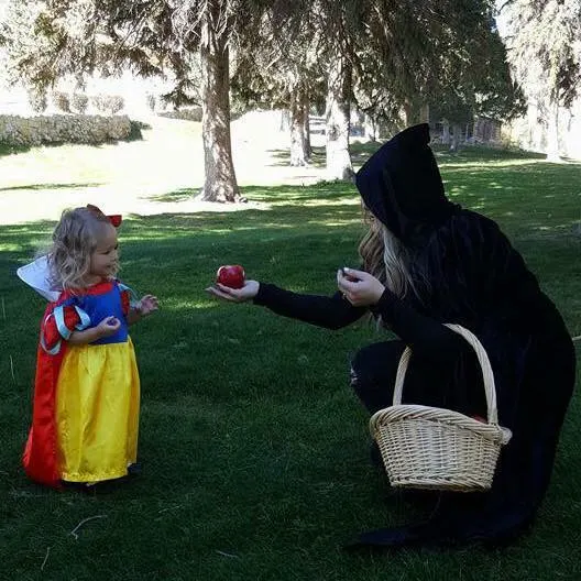 Woman wearing witch costume holds out apple to toddler girl wearing Snow White Halloween costume.