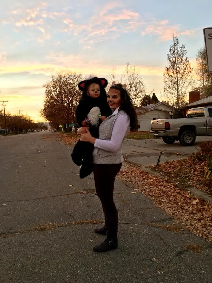 Woman wearing deer costume holds toddler boy wearing skunk Halloween costume.