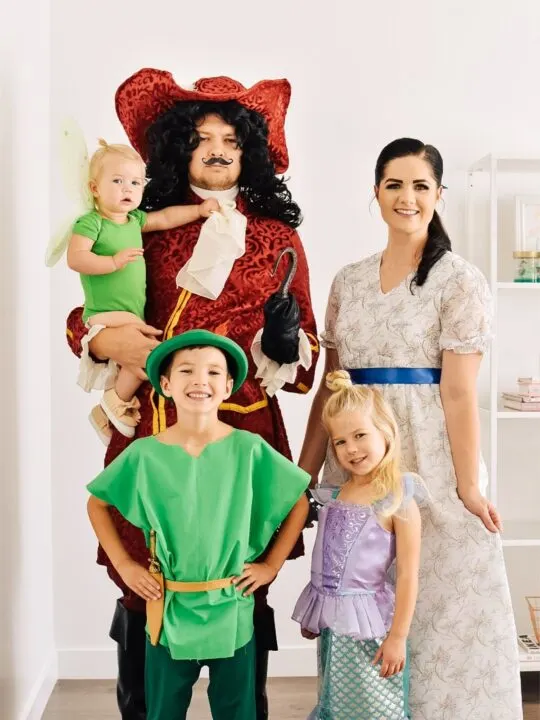 Family wearing Peter Pan character costumes smiles in front of white wall.