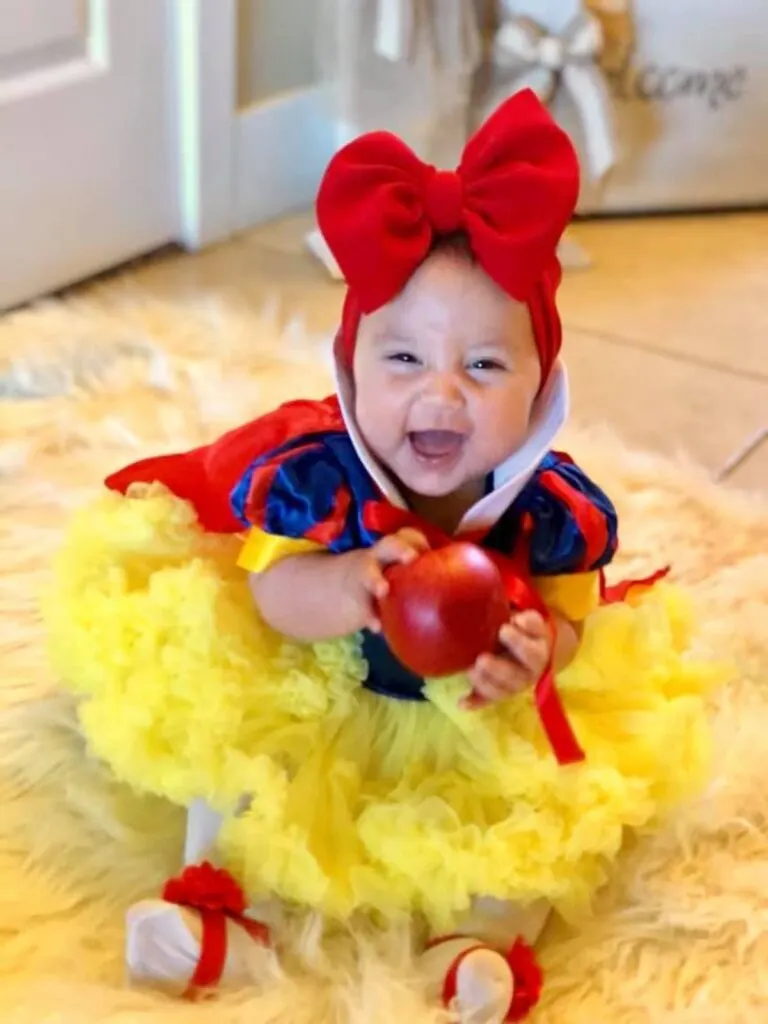 Baby wearing Snow White costume and holding apple smiles while sitting on floor.
