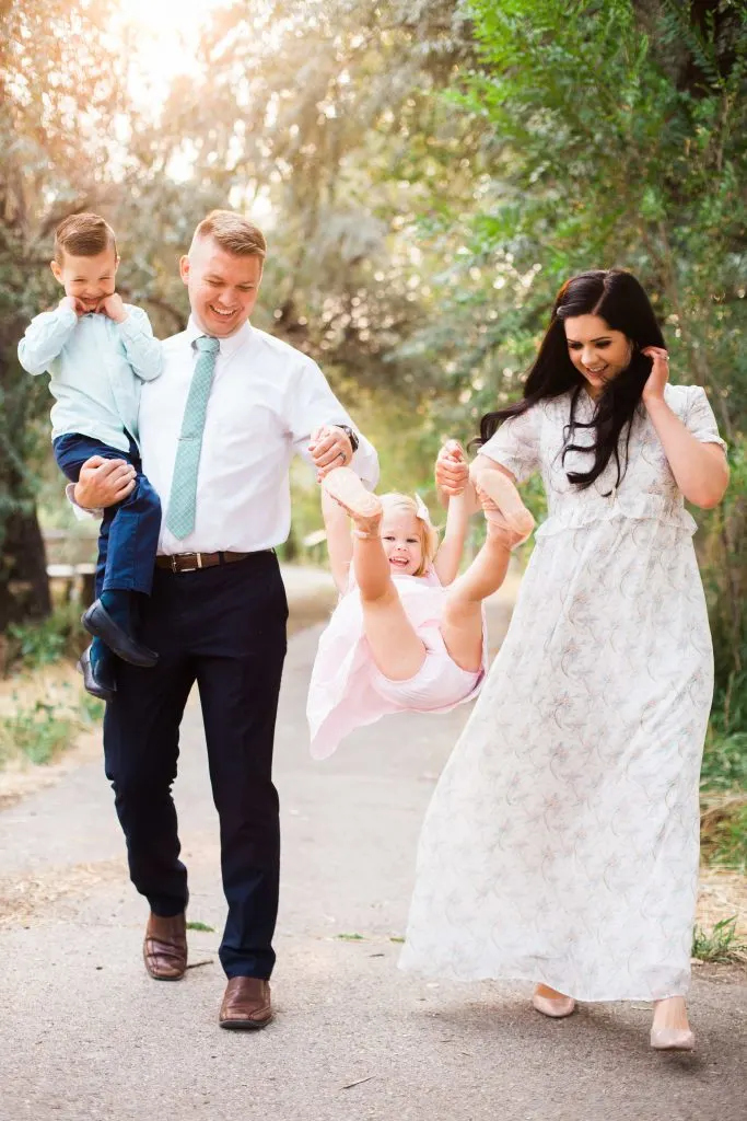 Man holding young boy and woman wearing floral dress swing toddler girl during family photos.