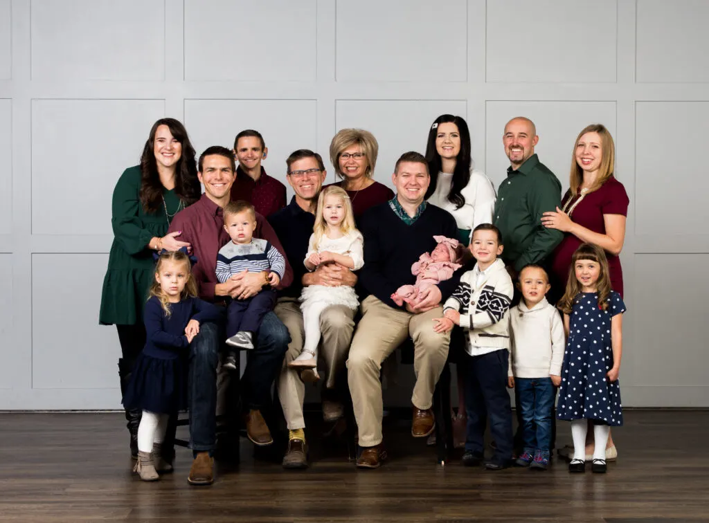 Large family of 16 wearing burgundy, green, and cream outfits smiles in front of gray wall.