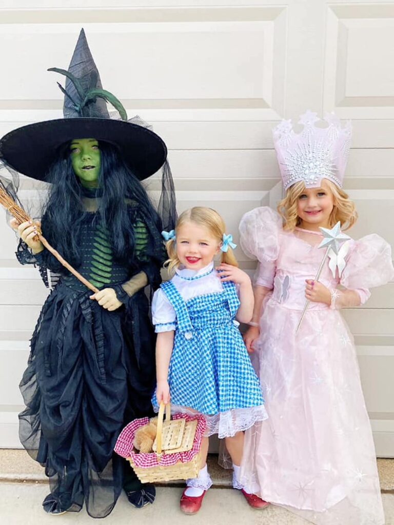 Three girls wearing Wizard of Oz character costumes smile in front of garage door.