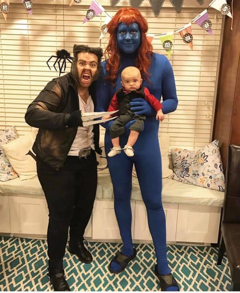 Family wearing X-Men character costumes smile in front of Halloween decorations.