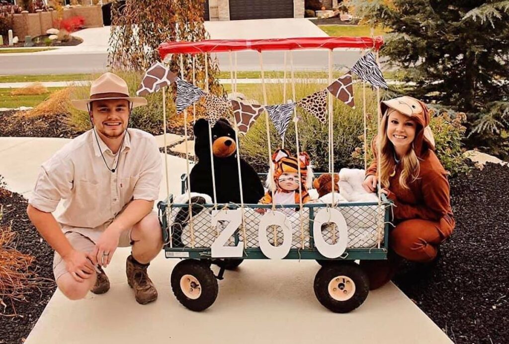 Man wearing zoo keeper costume and woman and baby wearing animal costumes smile next to DIY cage.