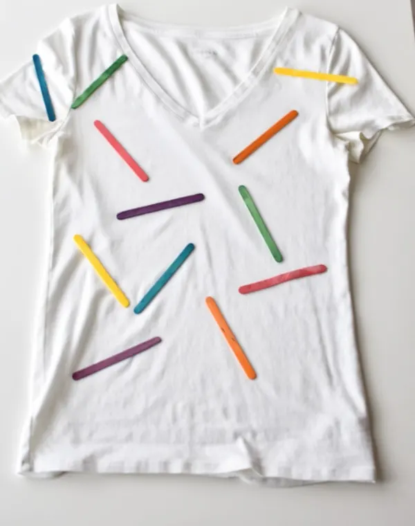 White t-shirt with rainbow colored popsicle sticks attached laying on white table.